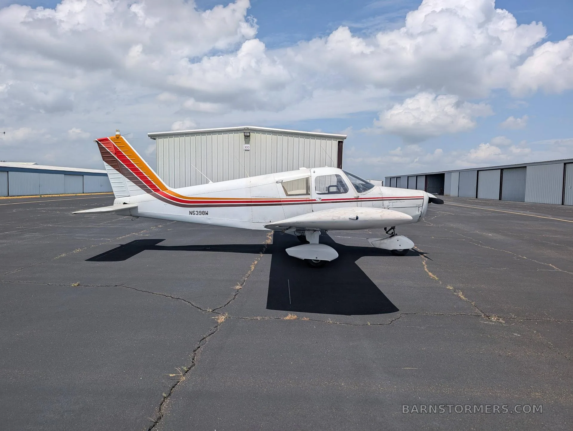A white single-engine low-wing aircraft with red, orange, and brown stripes on the tail and fuselage, viewed from the right side, parked on an asphalt ramp.