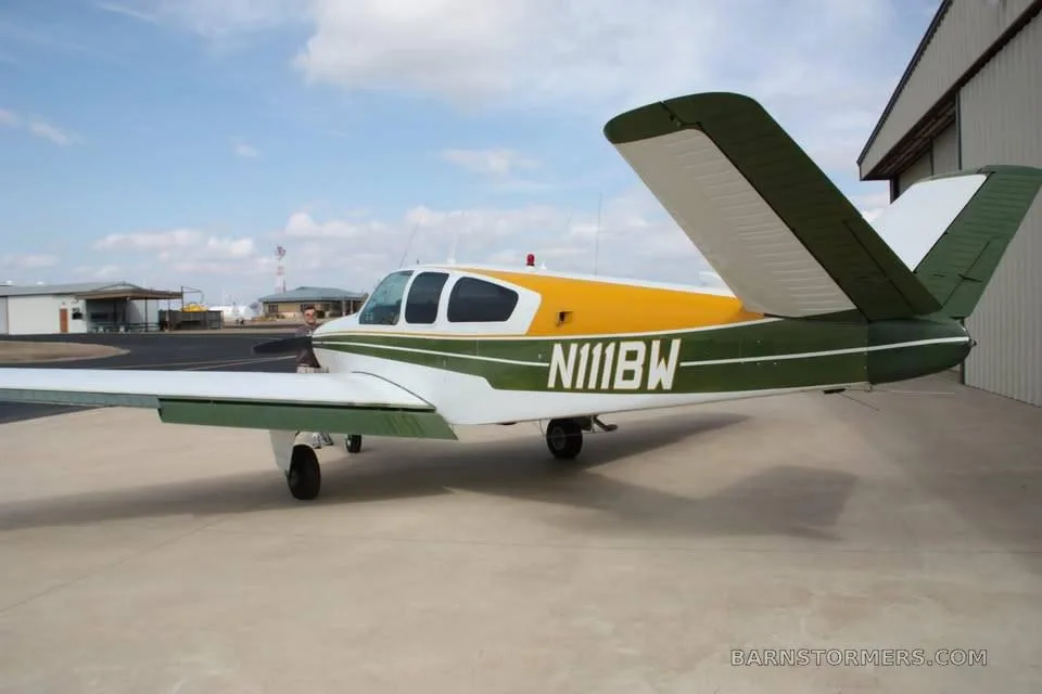 A green, white, and yellow single-engine propeller aircraft (N111BW) parked on an airfield next to a hangar.