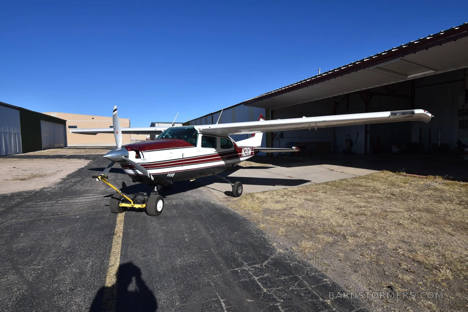 A white and maroon Cessna 210 aircraft with registration N210W is parked on asphalt next to an open hangar under a clear blue sky.