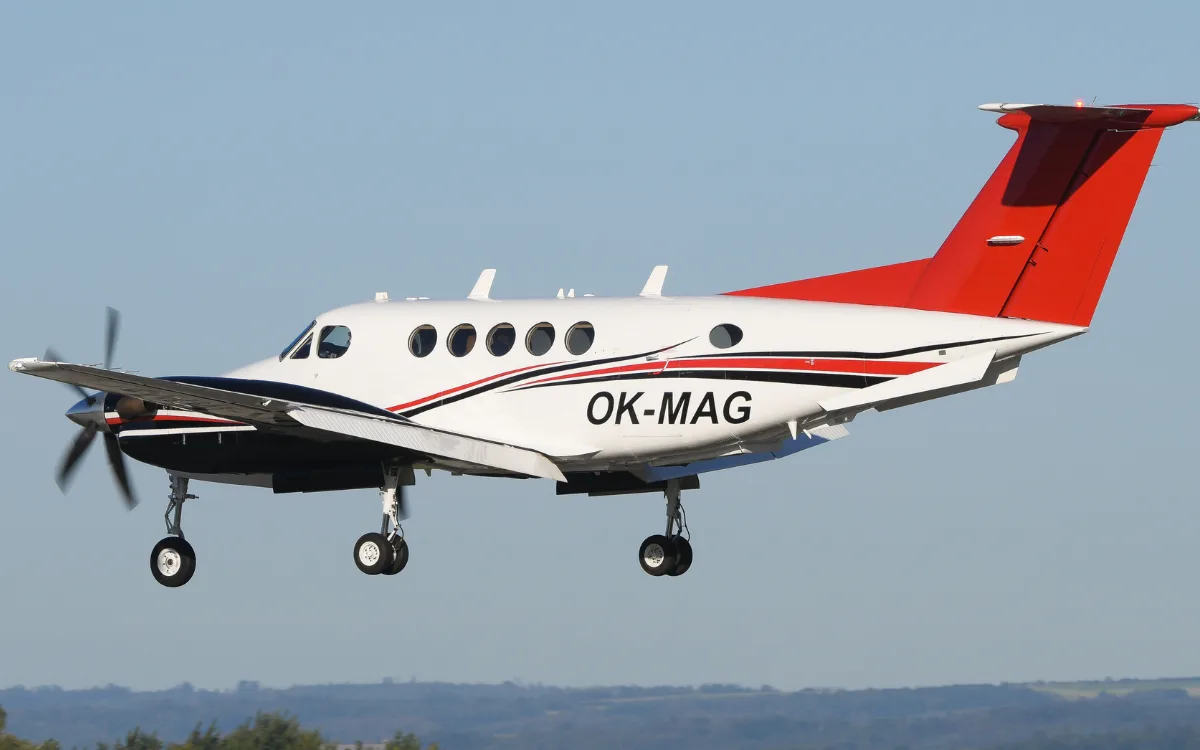 A white and black twin-turboprop aircraft with a red tail and registration OK-MAG is shown in flight with landing gear extended.