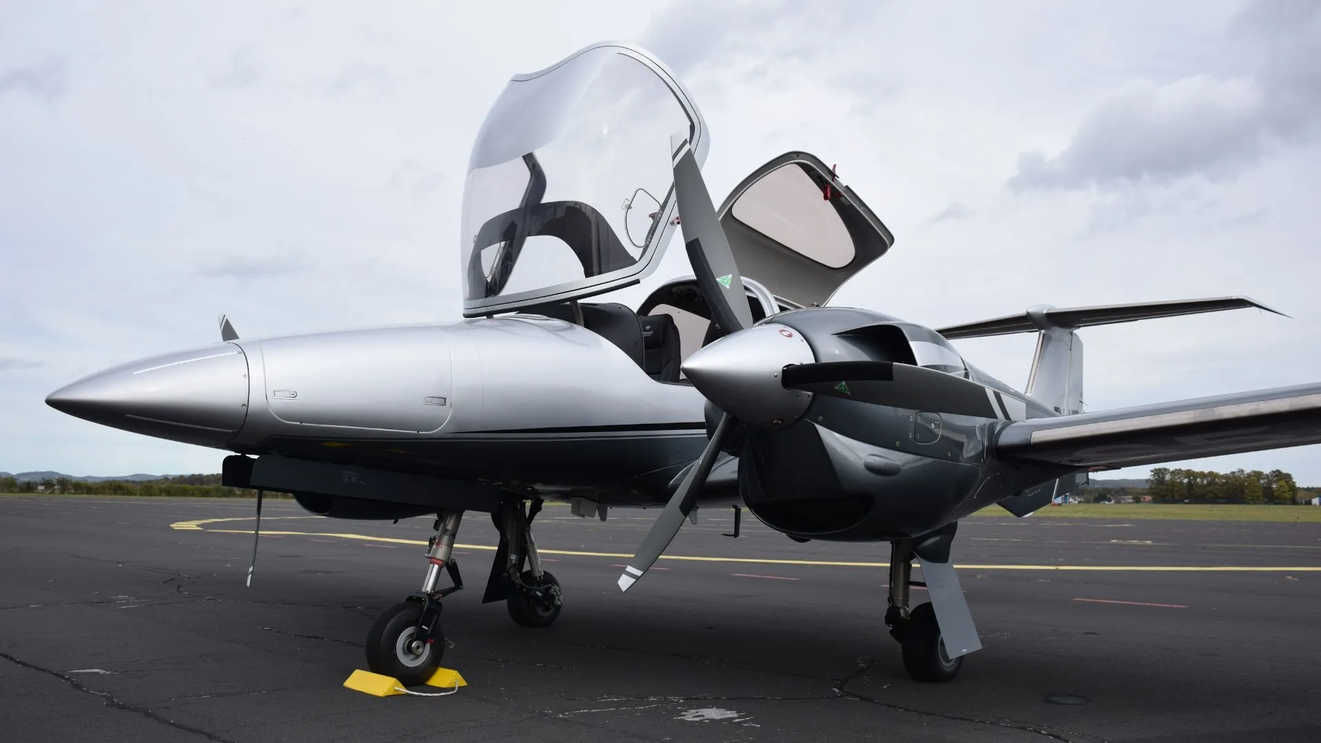 A three-quarter front view of a silver Diamond DA62 twin-engine aircraft with its canopy open on an airfield.