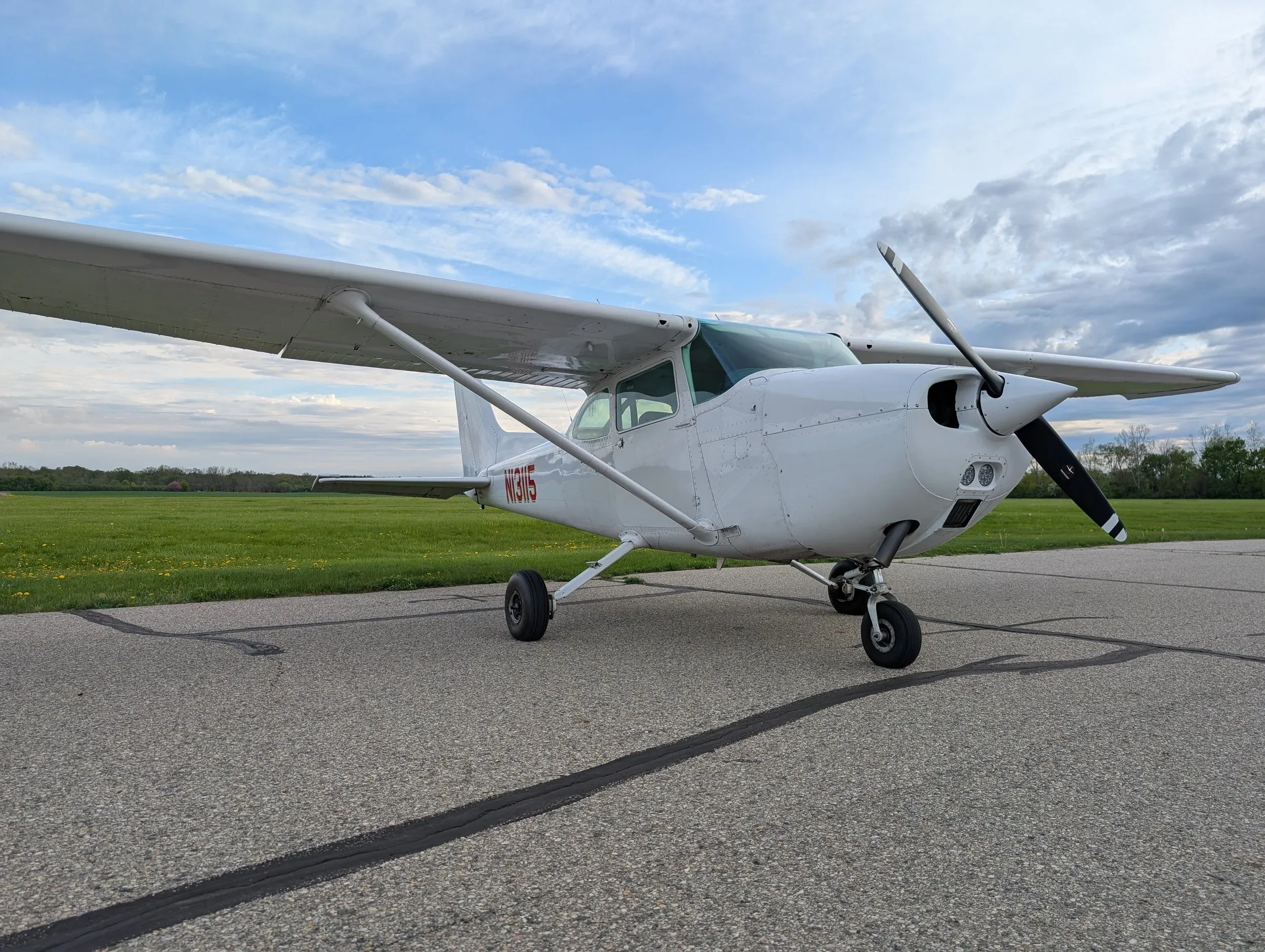 A white single-engine propeller aircraft, likely a Cessna, with registration N13115, parked on an asphalt ramp next to a green field under a cloudy sky.