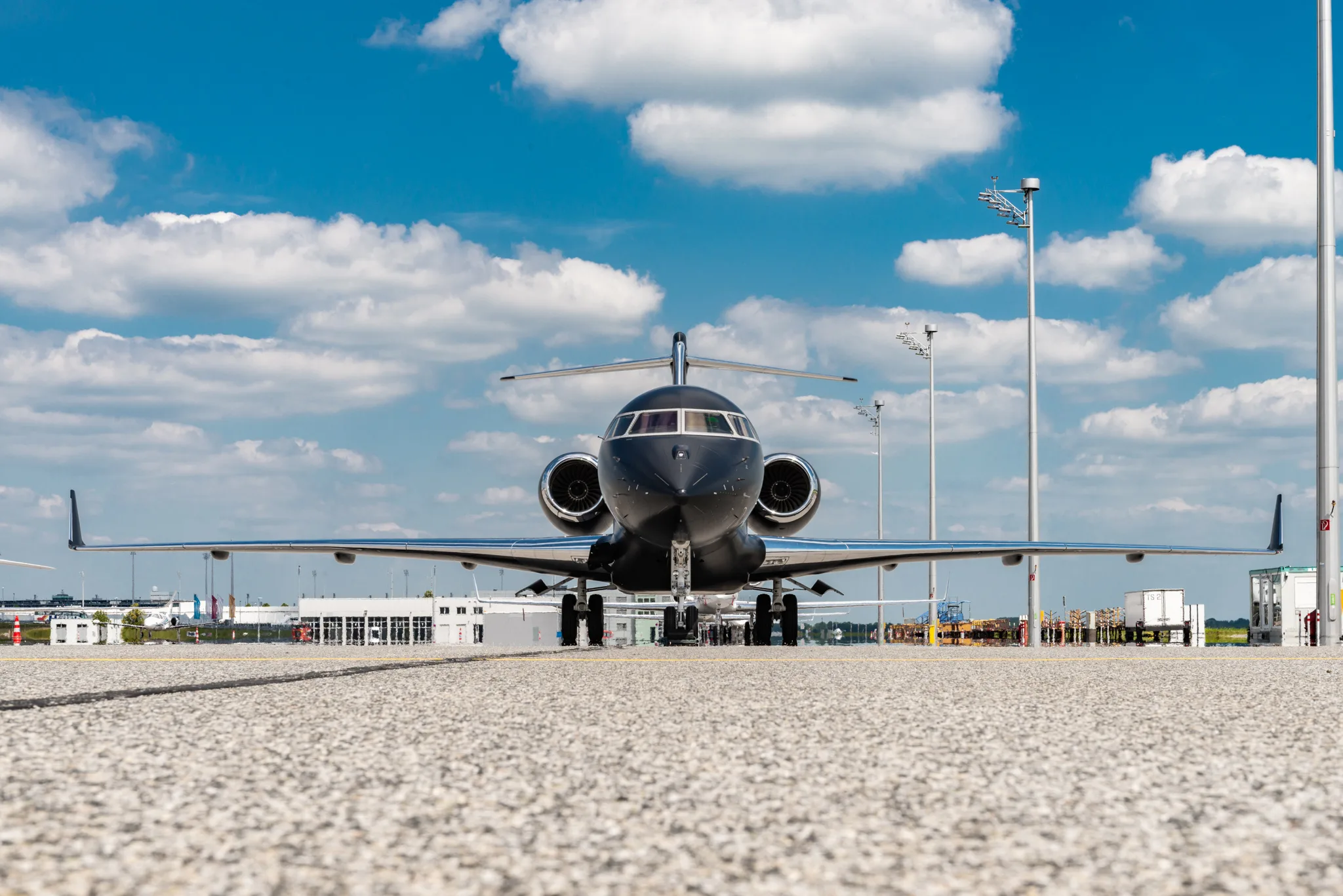A black business jet is seen head-on on an airport tarmac under a bright blue sky with white clouds.