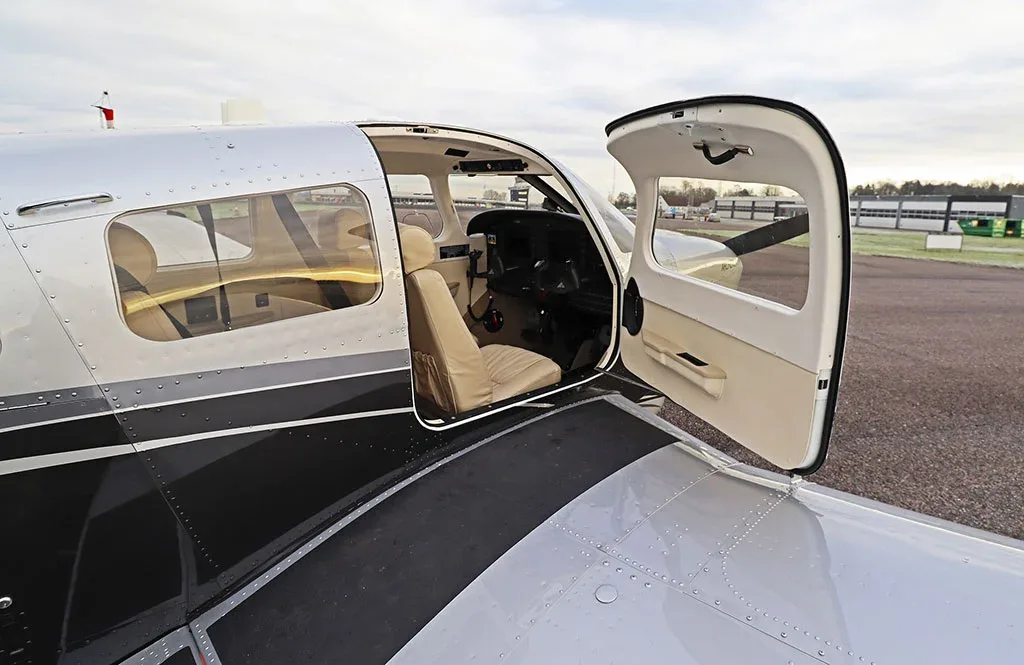 The open door of a silver and black Piper Archer LX aircraft reveals the light tan interior and cockpit.