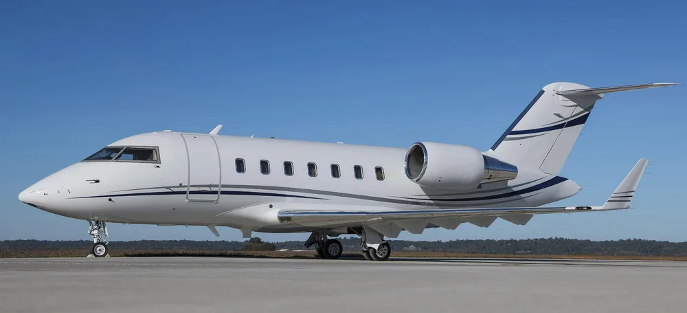 A white Bombardier Challenger 605 business jet with blue and grey stripes on the tail and fuselage, parked on an airfield under a clear blue sky.
