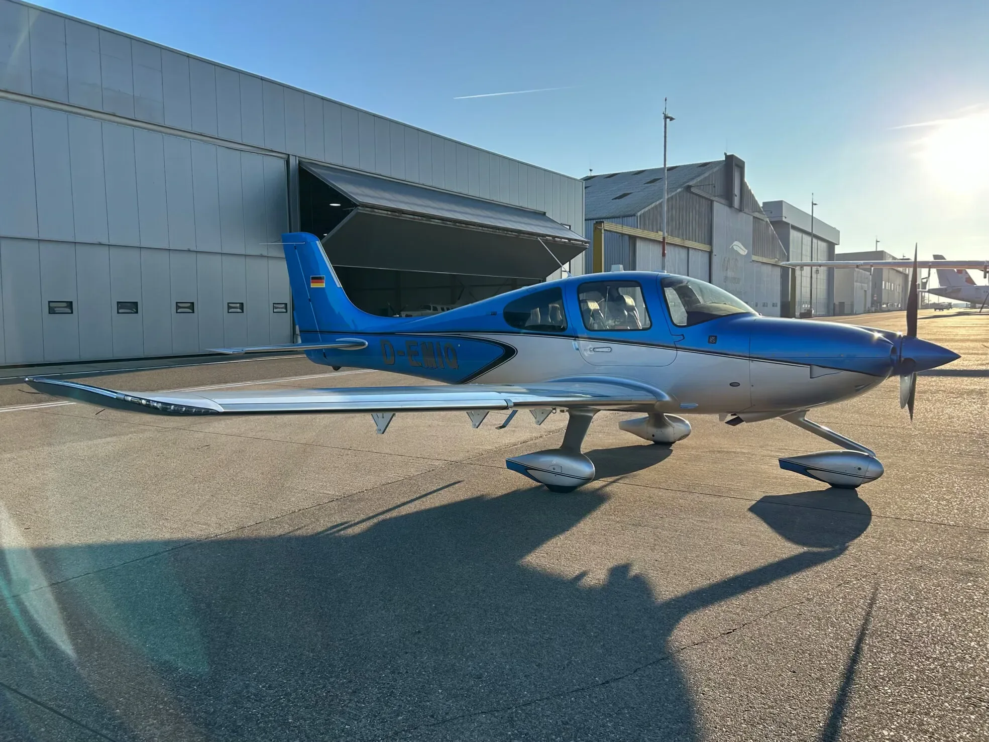 A blue and silver Cirrus SR22 aircraft parked on an asphalt apron with hangars in the background, bathed in late afternoon sun.