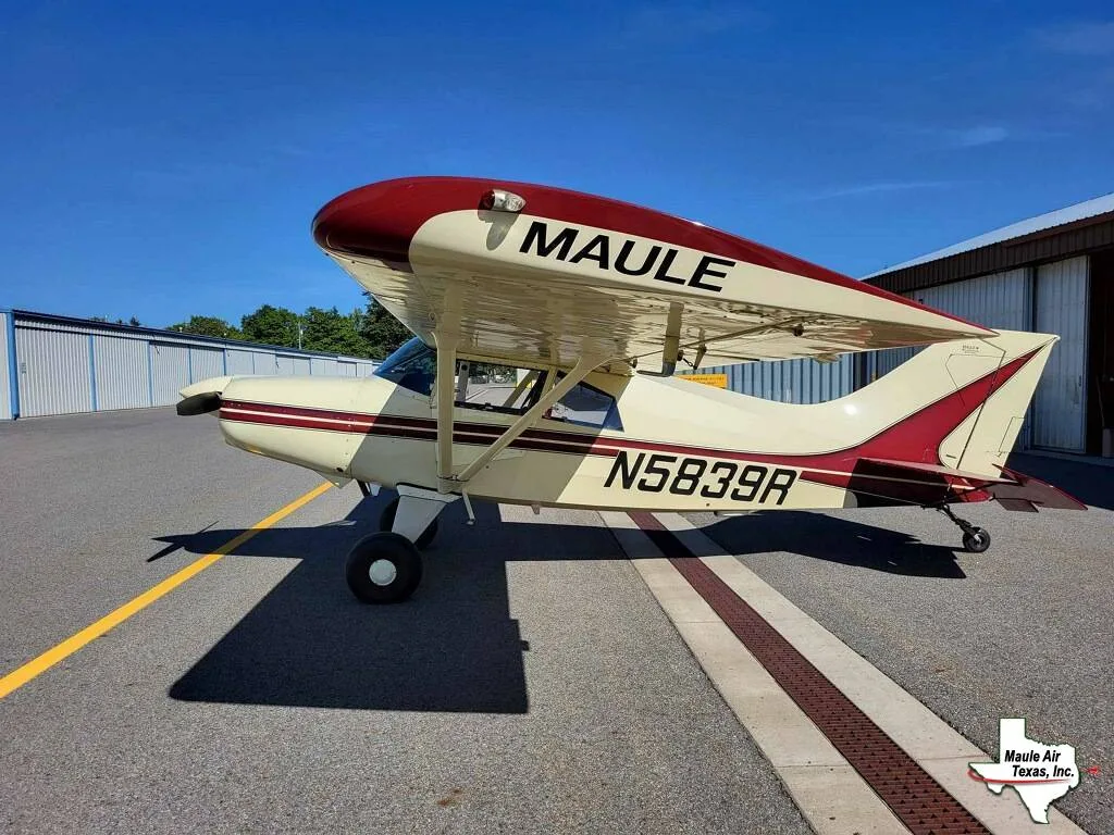 A cream and red Maule M-7-235B aircraft (N5839R) parked on an airfield tarmac with hangars in the background.