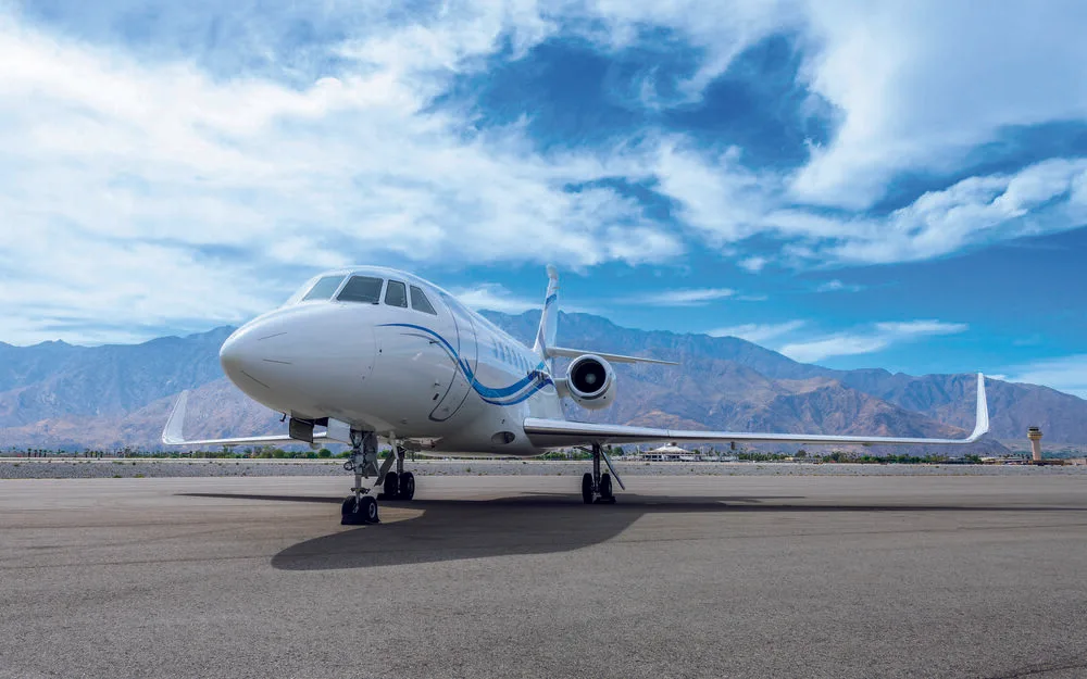 A front-quarter view of a white Dassault Falcon 2000LX business jet with blue stripes on the tarmac, with mountains and an air traffic control tower in the background.