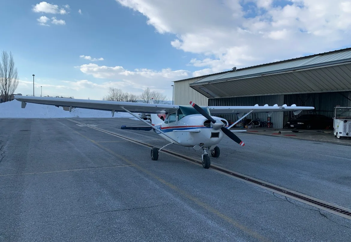 A white Cessna 182 with blue and red stripes parked on a snowy tarmac in front of a hangar.