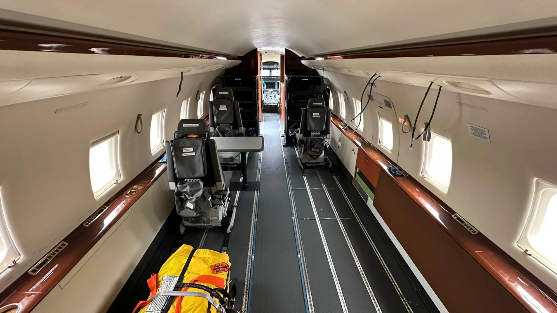 The interior cabin of an aircraft configured with tracks on the floor, several seats, and a yellow stretcher, looking towards the cockpit.
