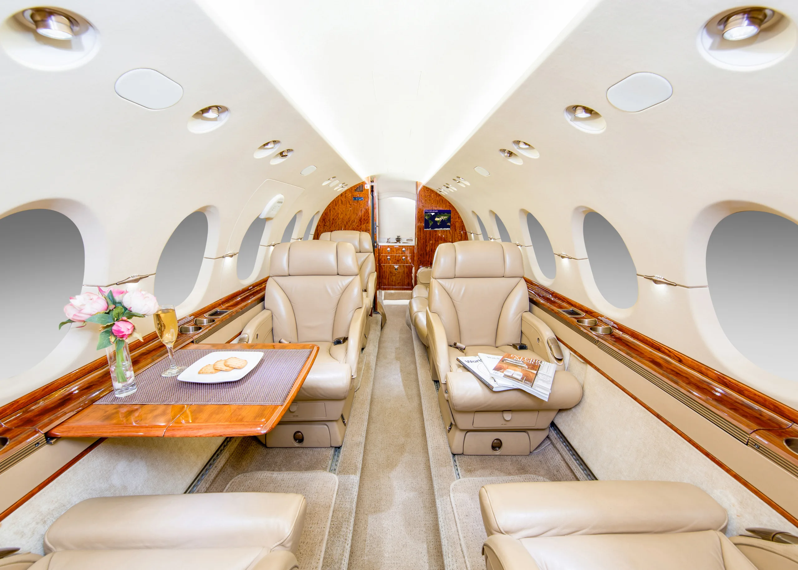 The luxurious interior cabin of a private jet, featuring beige leather seats, a polished wood table with flowers and snacks, and large oval windows.