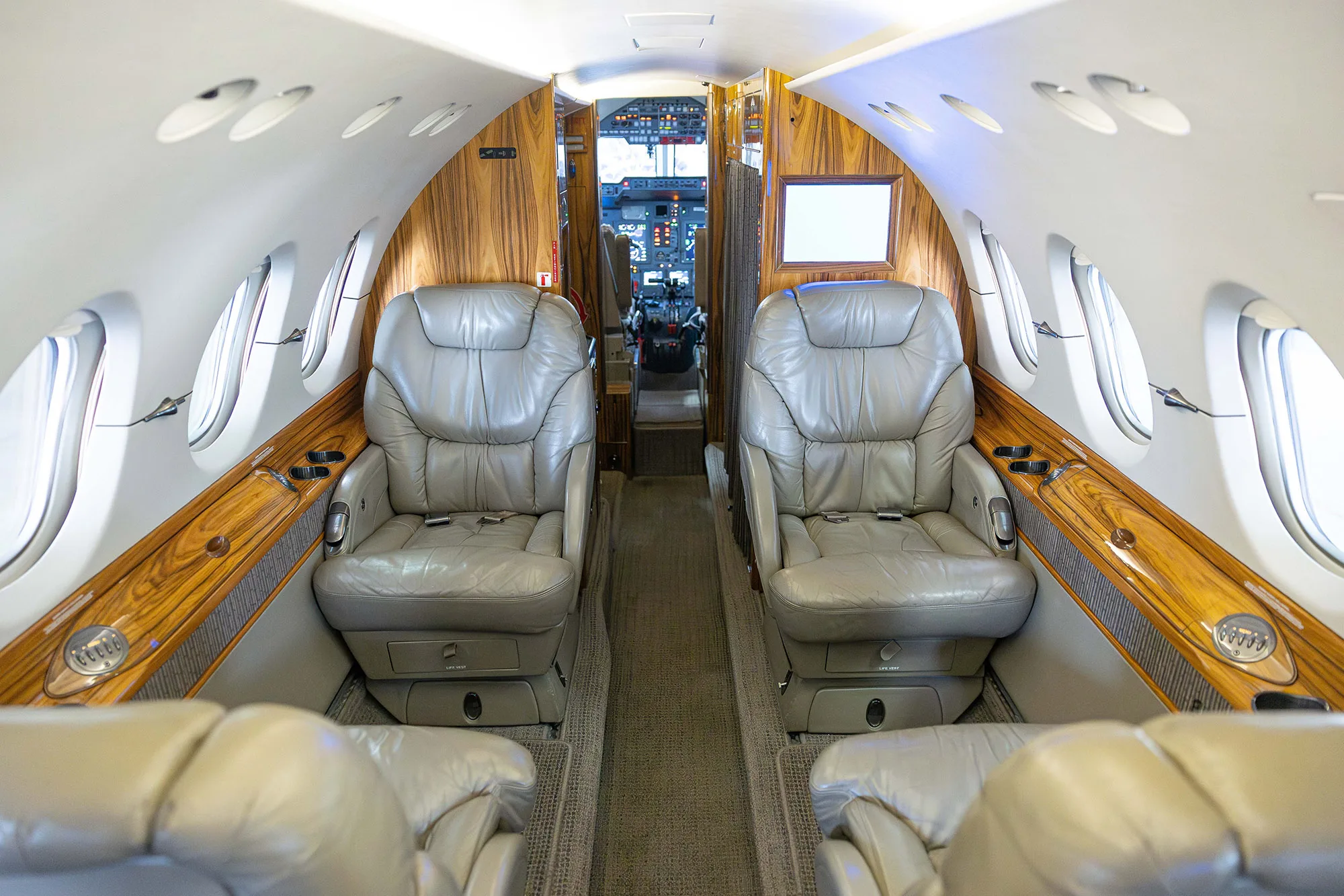 The interior cabin of a business jet with grey leather seats and wood paneling, looking towards the cockpit.