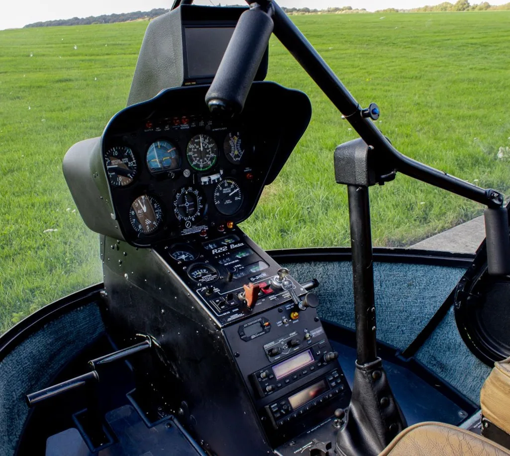The instrument panel and controls of a Robinson R22 Beta helicopter with a view of a green field outside.