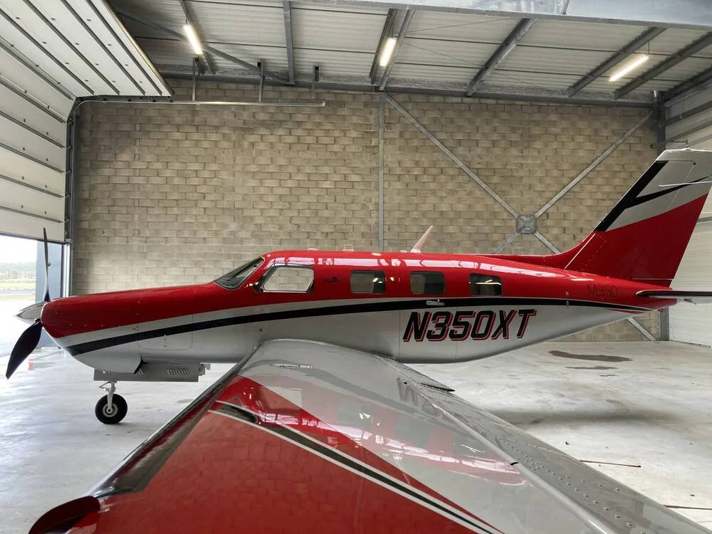 A side view of the red and silver Piper M350 aircraft, N350XT, with its wing in the foreground, inside a hangar.