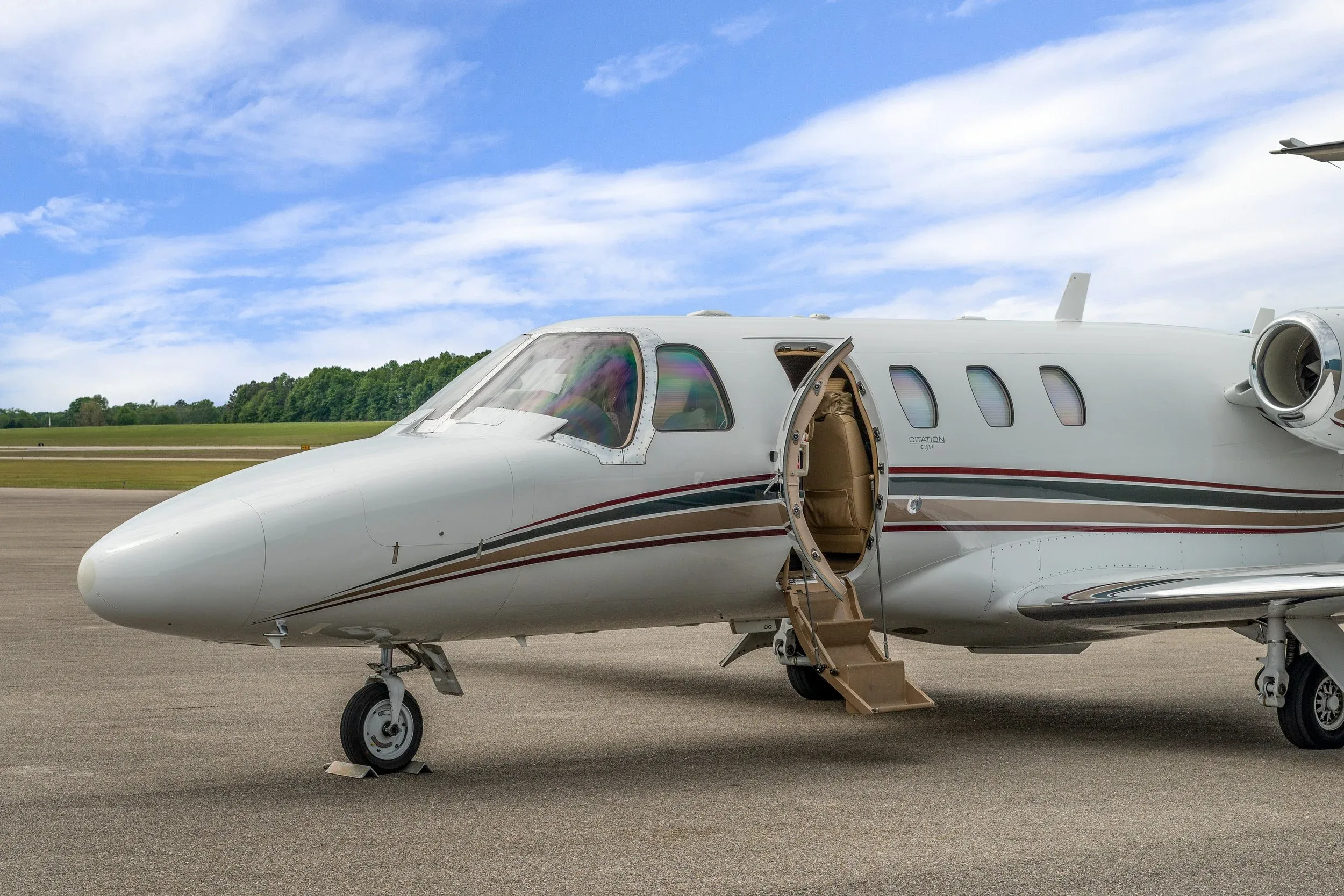 A white Cessna Citation CJ1+ private jet with brown and red stripes sits on an airport tarmac with its main door open and stairs extended.