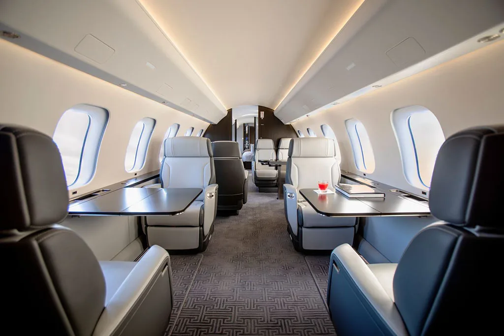 The luxurious interior cabin of a private jet, featuring light grey and black leather seats with tables, and a patterned carpet.