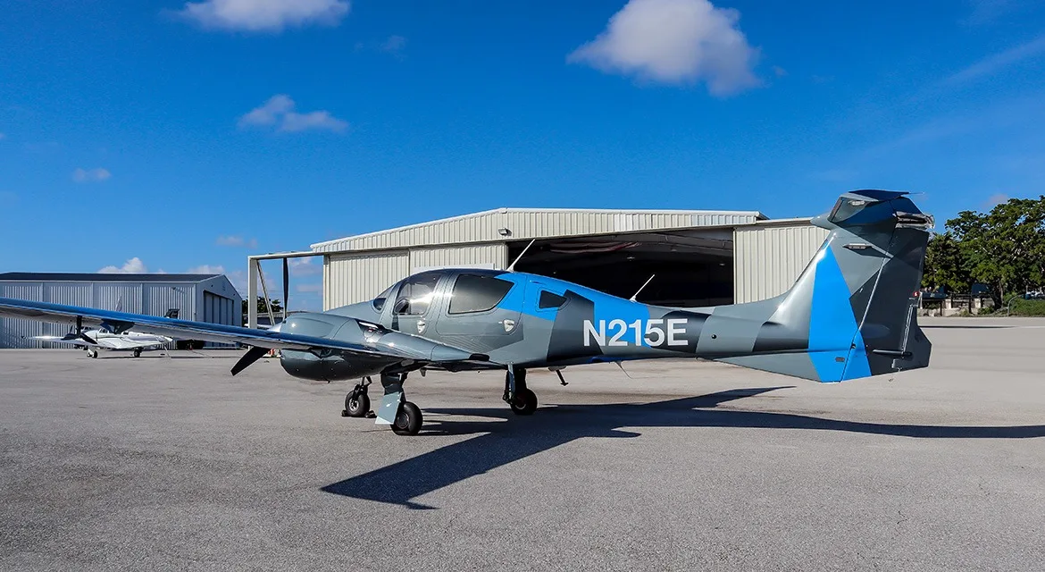 A gray and blue Diamond DA62 aircraft, registration N215E, parked on an airfield tarmac next to a hangar.