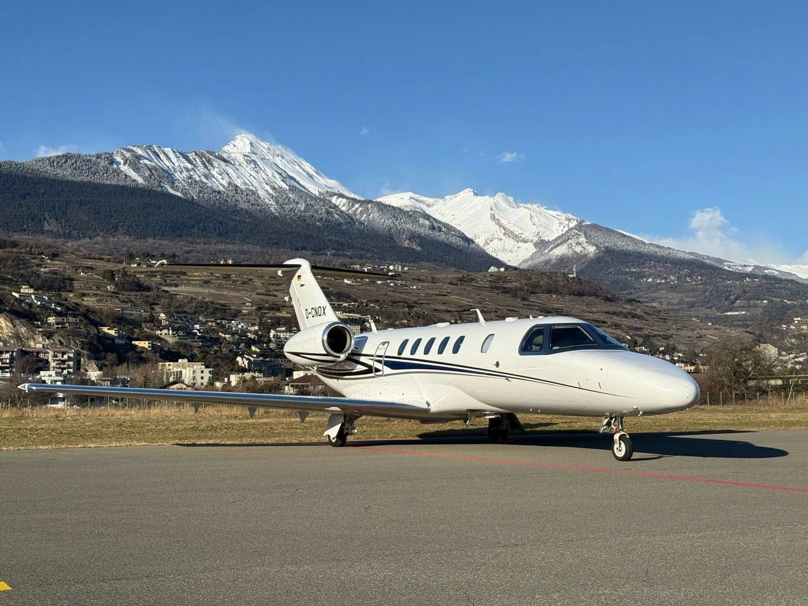 A white Cessna CitationJet with dark blue and black stripes is parked on an asphalt runway, with snow-capped mountains and a valley town in the background under a clear blue sky.