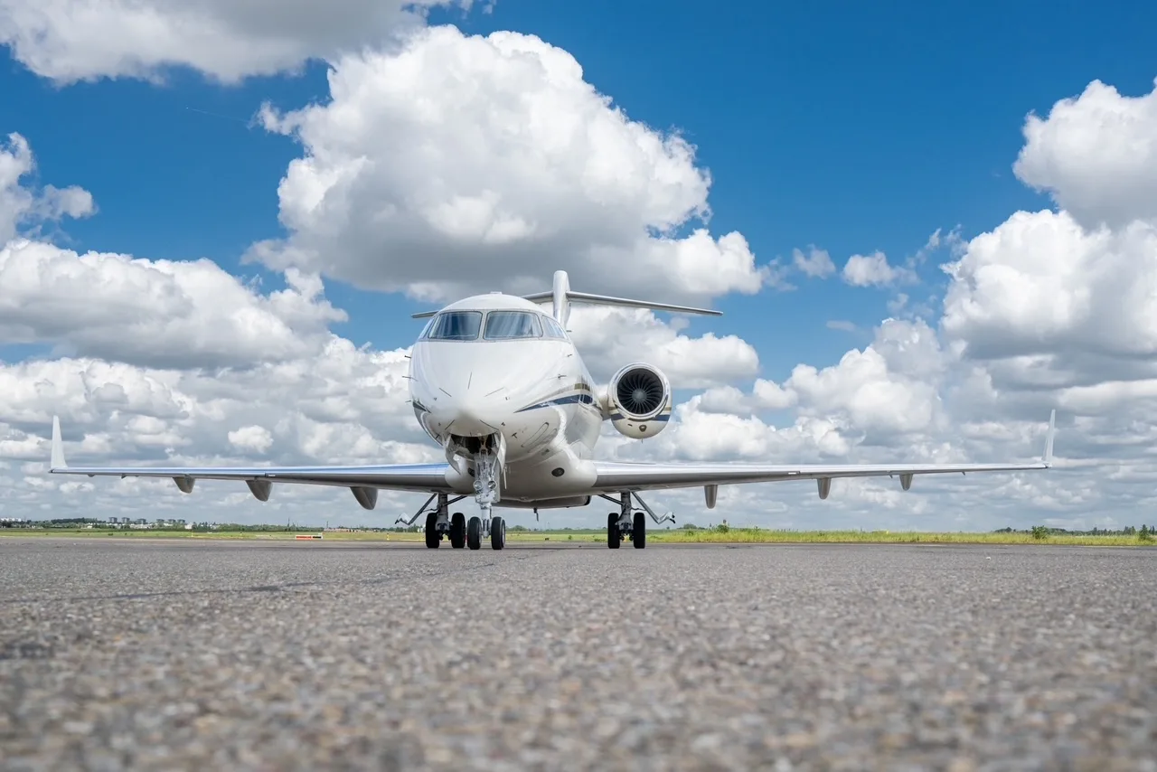 A white Bombardier Challenger 300 business jet seen from the front on an airfield under a blue sky with clouds.