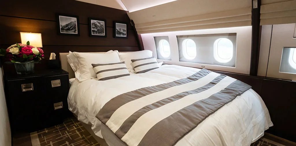 A luxurious aircraft bedroom with a large bed, dark wood nightstand, and three small windows.