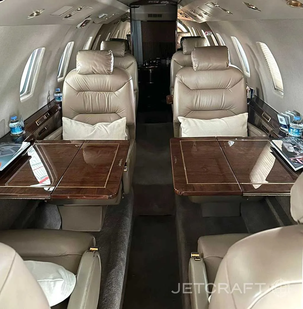 The interior cabin of a private jet with tan leather seats and polished wood tables deployed, showing magazines and water bottles.