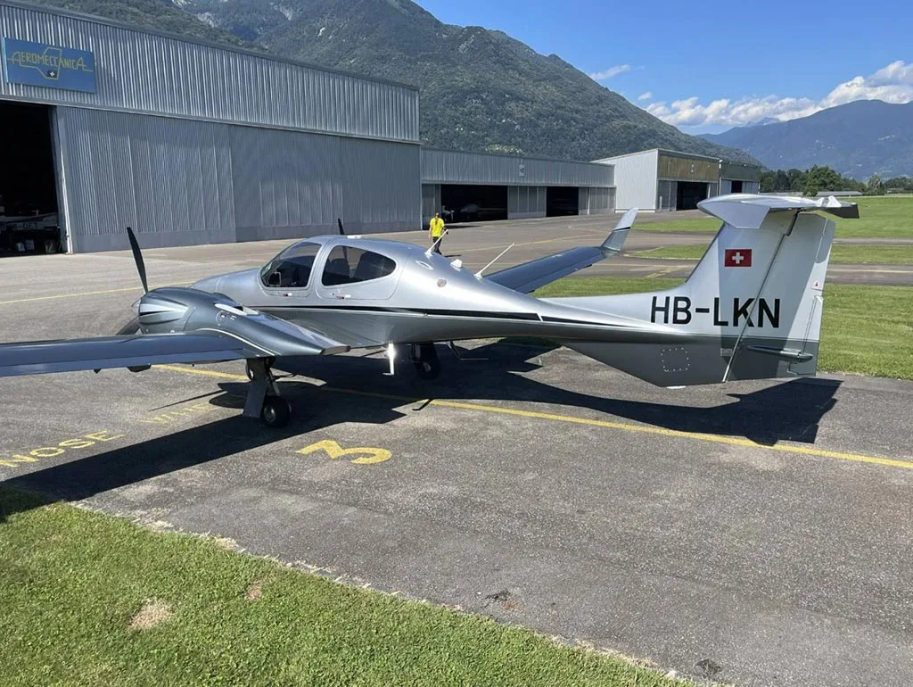 A silver Diamond DA42 aircraft with registration HB-LKN parked on an airfield next to a hangar with mountains in the background.