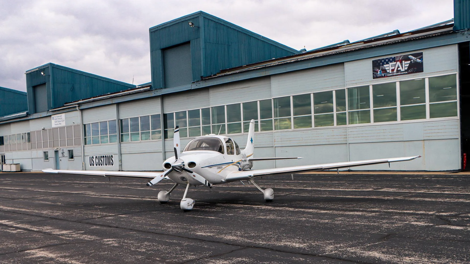 A white Cirrus aircraft with blue and gold stripes is parked on a tarmac in front of a hangar with a 'US CUSTOMS' sign.