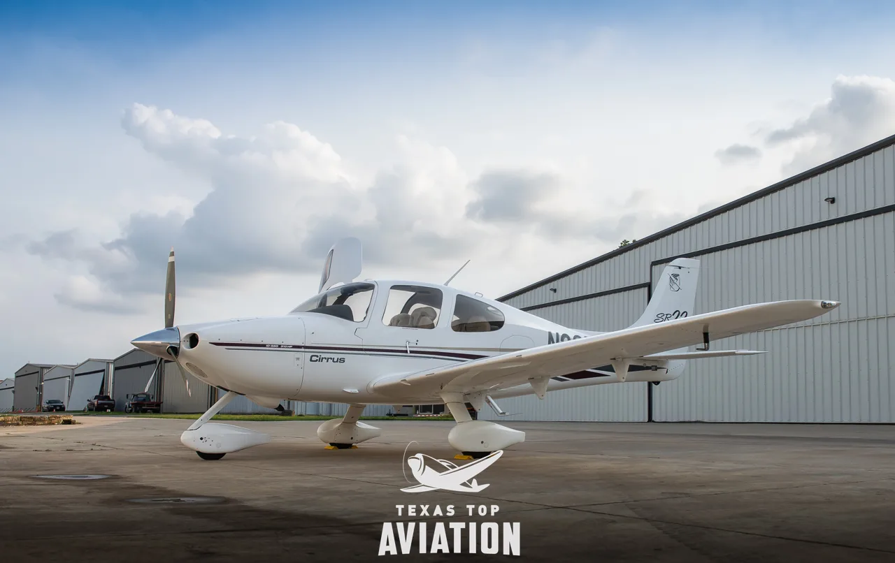 A white Cirrus SR22 aircraft with maroon and gold stripes is parked on a tarmac in front of hangars under a partly cloudy sky.
