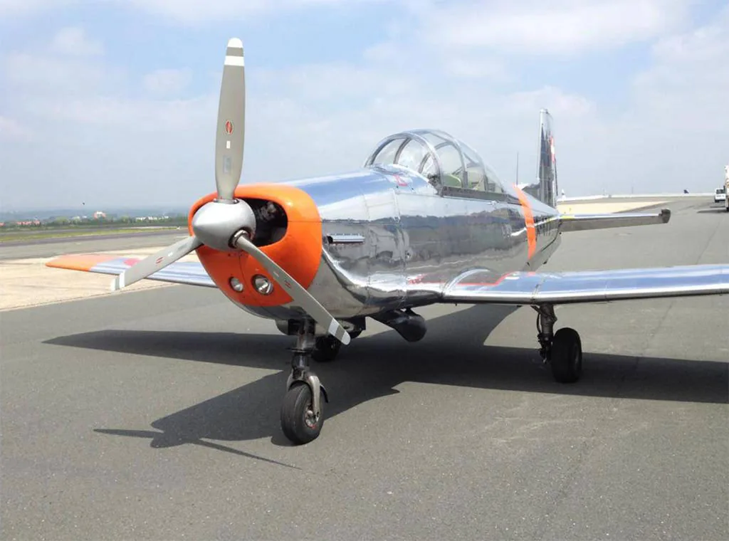 A front-quarter view of a polished silver and orange Pilatus P-3 aircraft with a propeller, parked on an airfield.