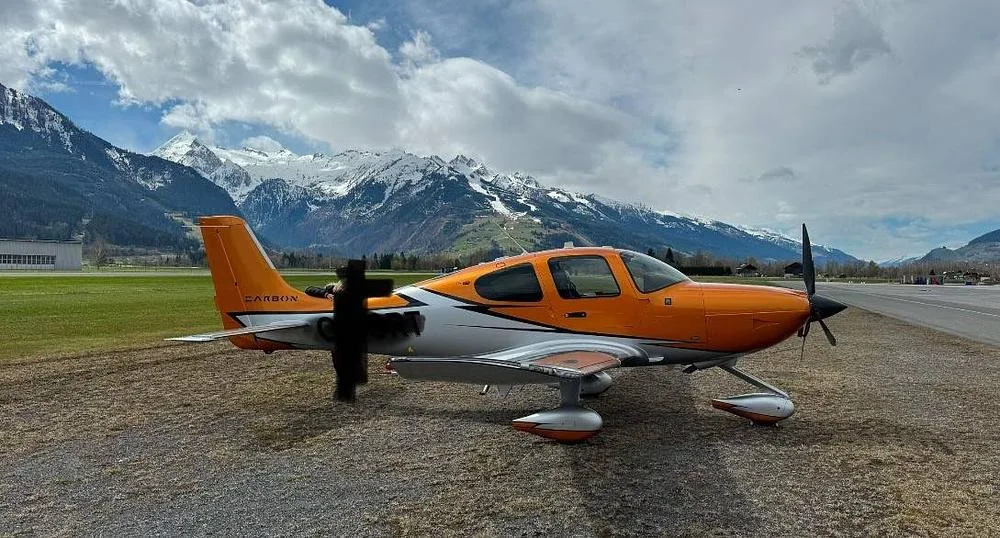 An orange and silver Cirrus SR22 aircraft parked on a grassy airfield with majestic snow-capped mountains in the background under a cloudy sky.