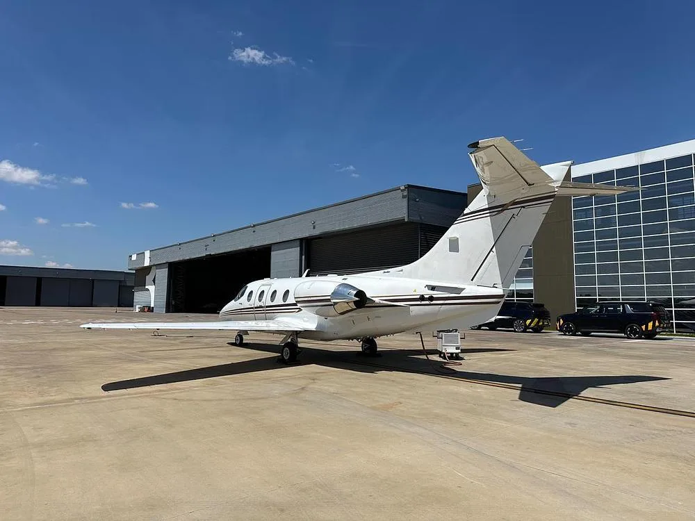 A white business jet with brown stripes seen from the rear-right, parked on a tarmac next to a hangar.