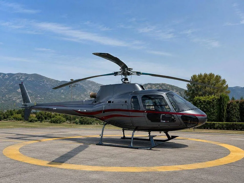 A gray helicopter with red stripes sits on a helipad with mountains and trees in the background under a blue sky.