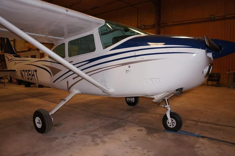 A white and blue Cessna 182 Skylane N735HT is parked in a hangar, viewed from the front-right.