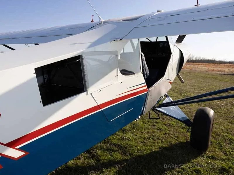 A close-up view of the white and blue Piper Super Cub's fuselage with an open door, parked on a grassy field.