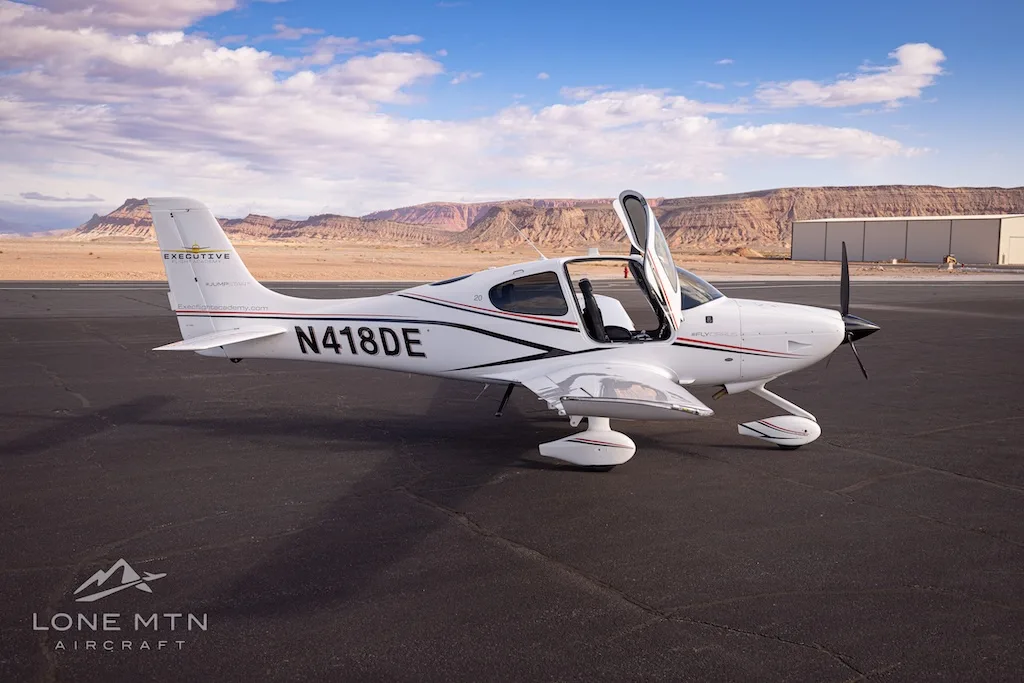 A white Cirrus SR22 aircraft with black and red stripes, registration N418DE, parked on an airfield with doors open.