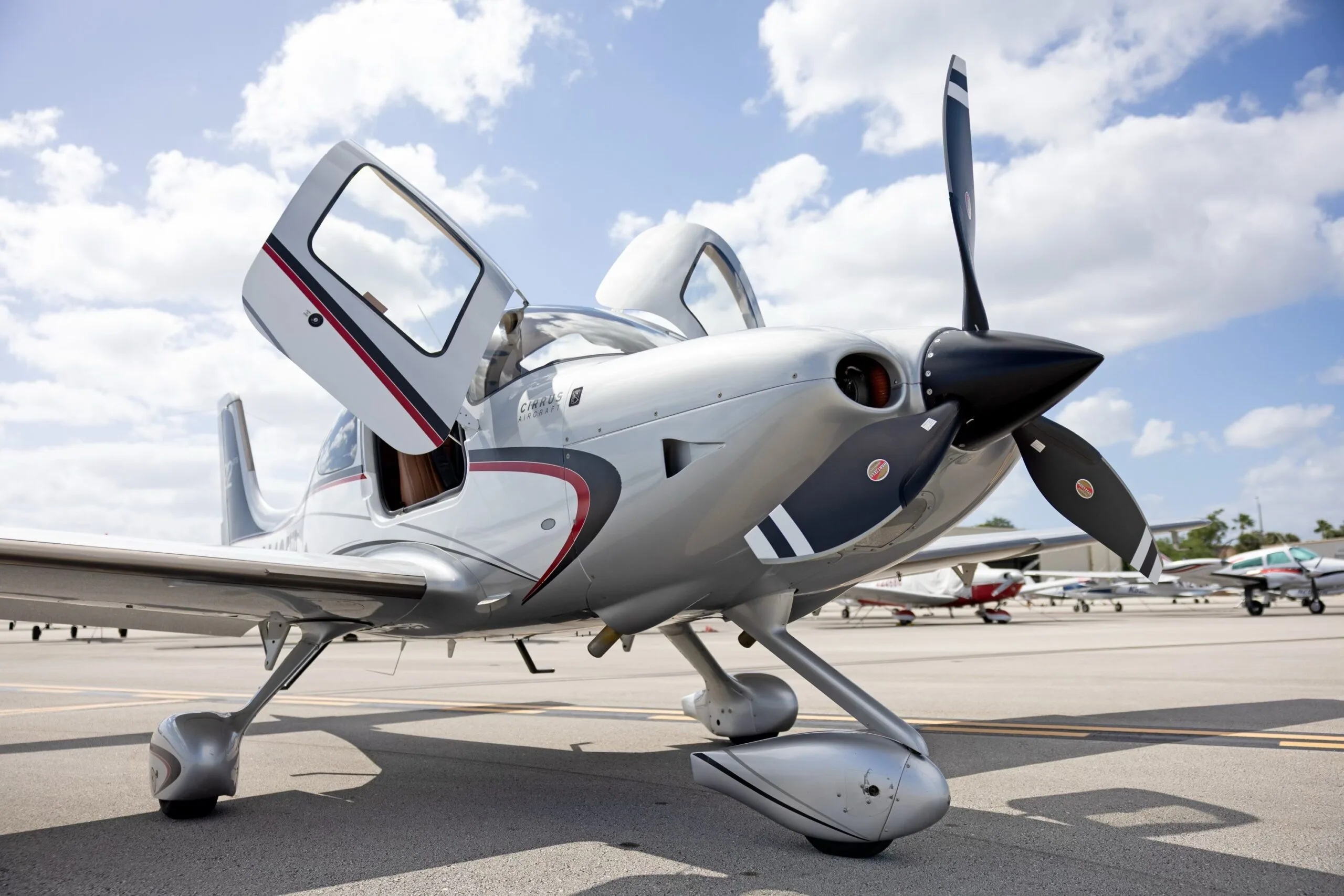 A front-quarter view of a silver Cirrus SR22 GTS aircraft with doors open, parked on an airfield with other planes in the background.