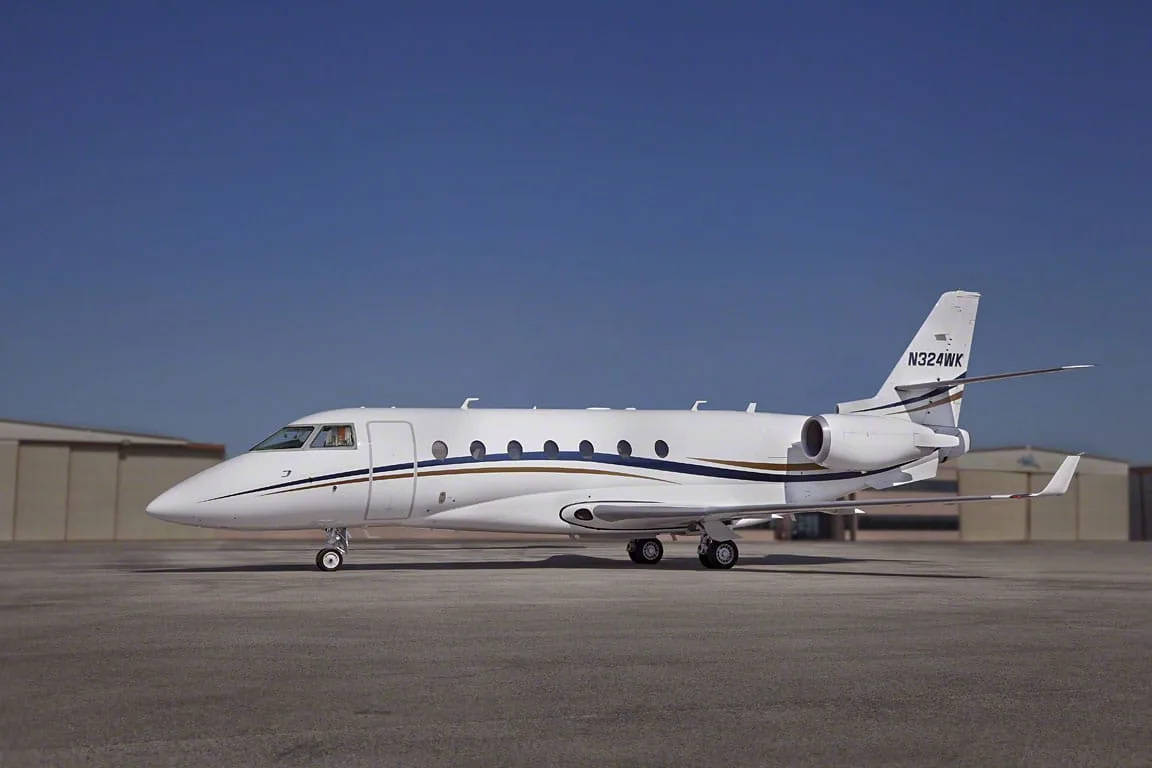 A white business jet with blue and gold stripes and tail number N324WK parked on a tarmac under a clear blue sky.