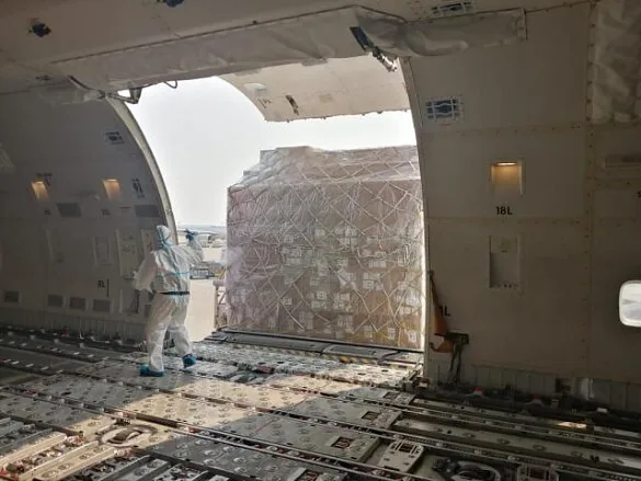 A person in a hazmat suit loads large, netted cargo into the open nose of a Boeing 747 cargo aircraft.