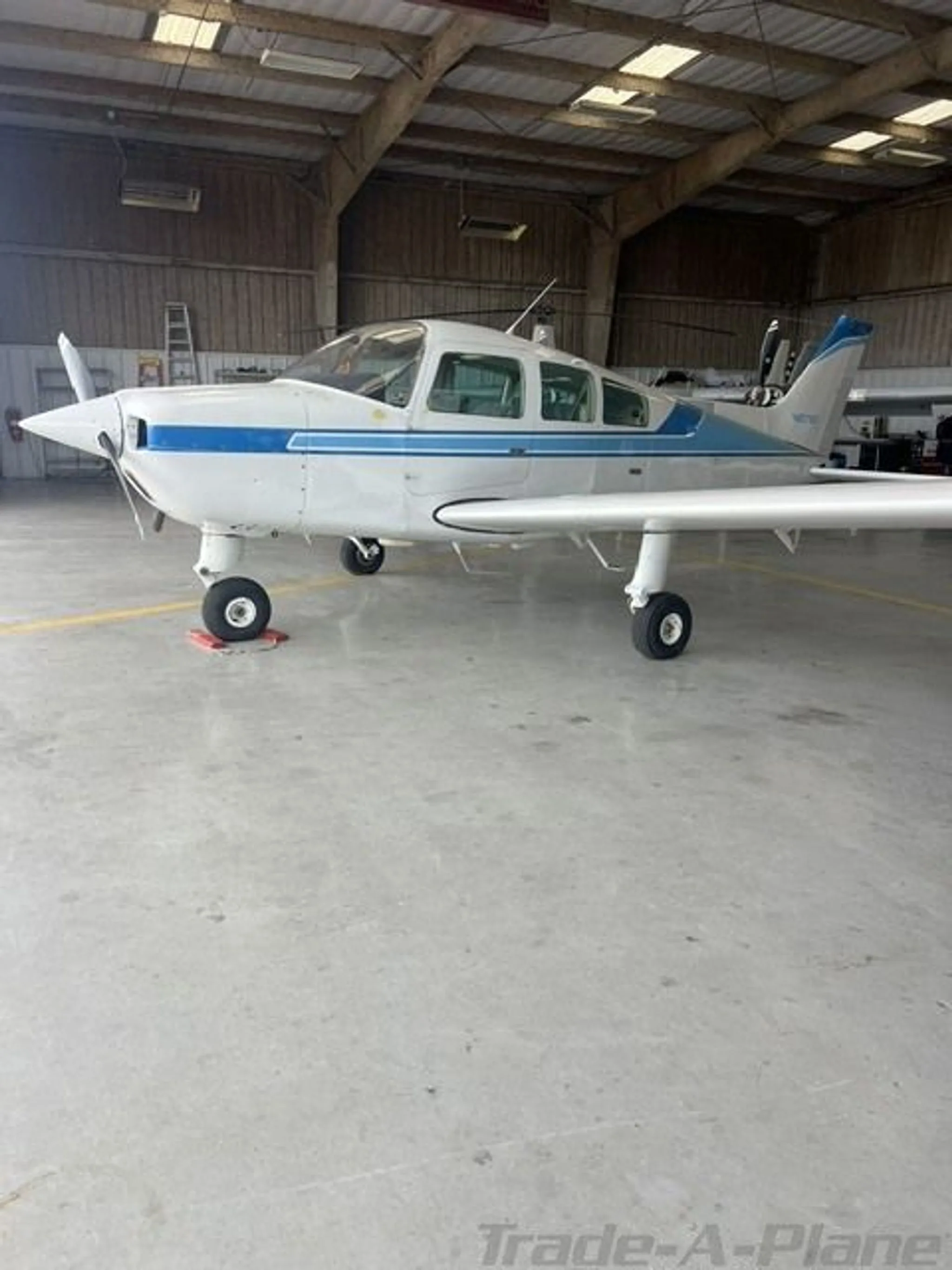 A white Beechcraft Sundowner N6718T with blue stripes parked inside a hangar, viewed from the side.