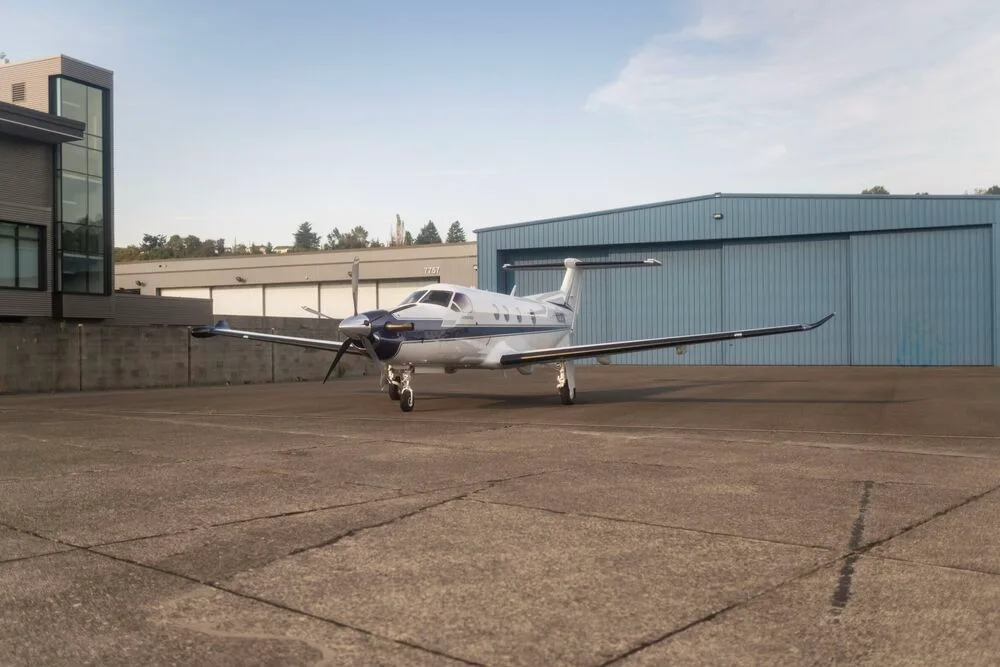 A white and blue Pilatus PC-12 aircraft with registration N226N parked on an tarmac in front of hangars.
