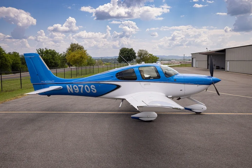 A side view of the blue and white Cirrus SR22 aircraft, N970S, parked on an asphalt ramp with a hangar in the background.