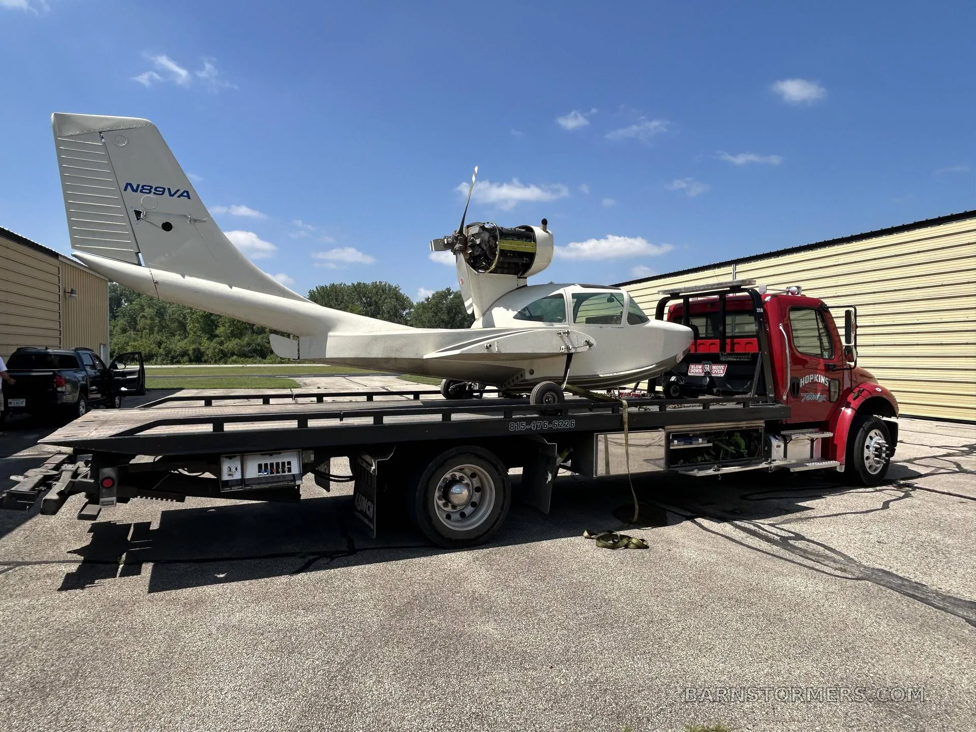 A white single-engine aircraft with tail number N89VA is loaded onto a red flatbed tow truck outdoors.