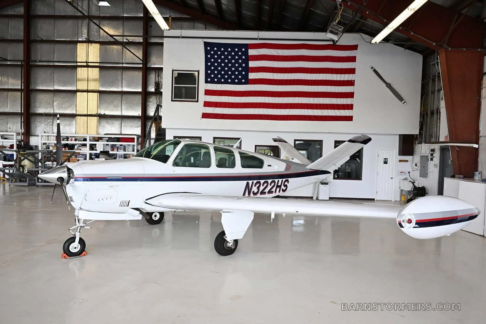 A white single-engine aircraft with blue and red stripes, registration N322HS, parked in a hangar with an American flag.
