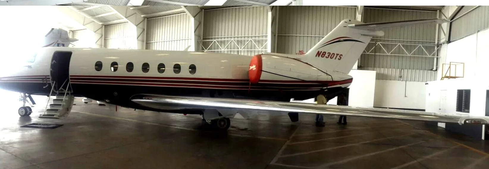 A white business jet with red and black stripes (N830TS) is parked inside a hangar with its main door open.