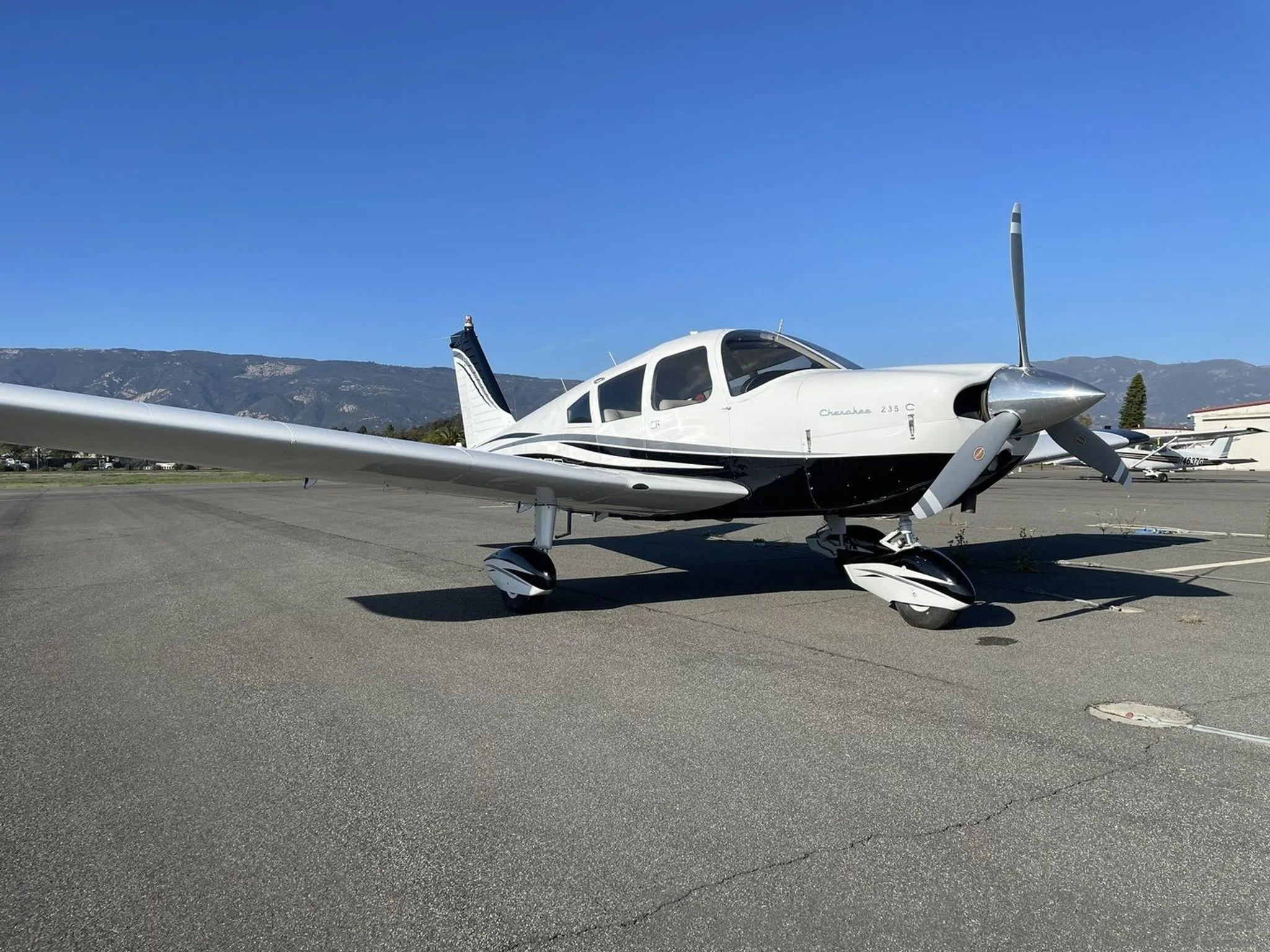 A white and black Piper Cherokee 235 C aircraft with registration N991CB parked on an airfield with mountains in the background.