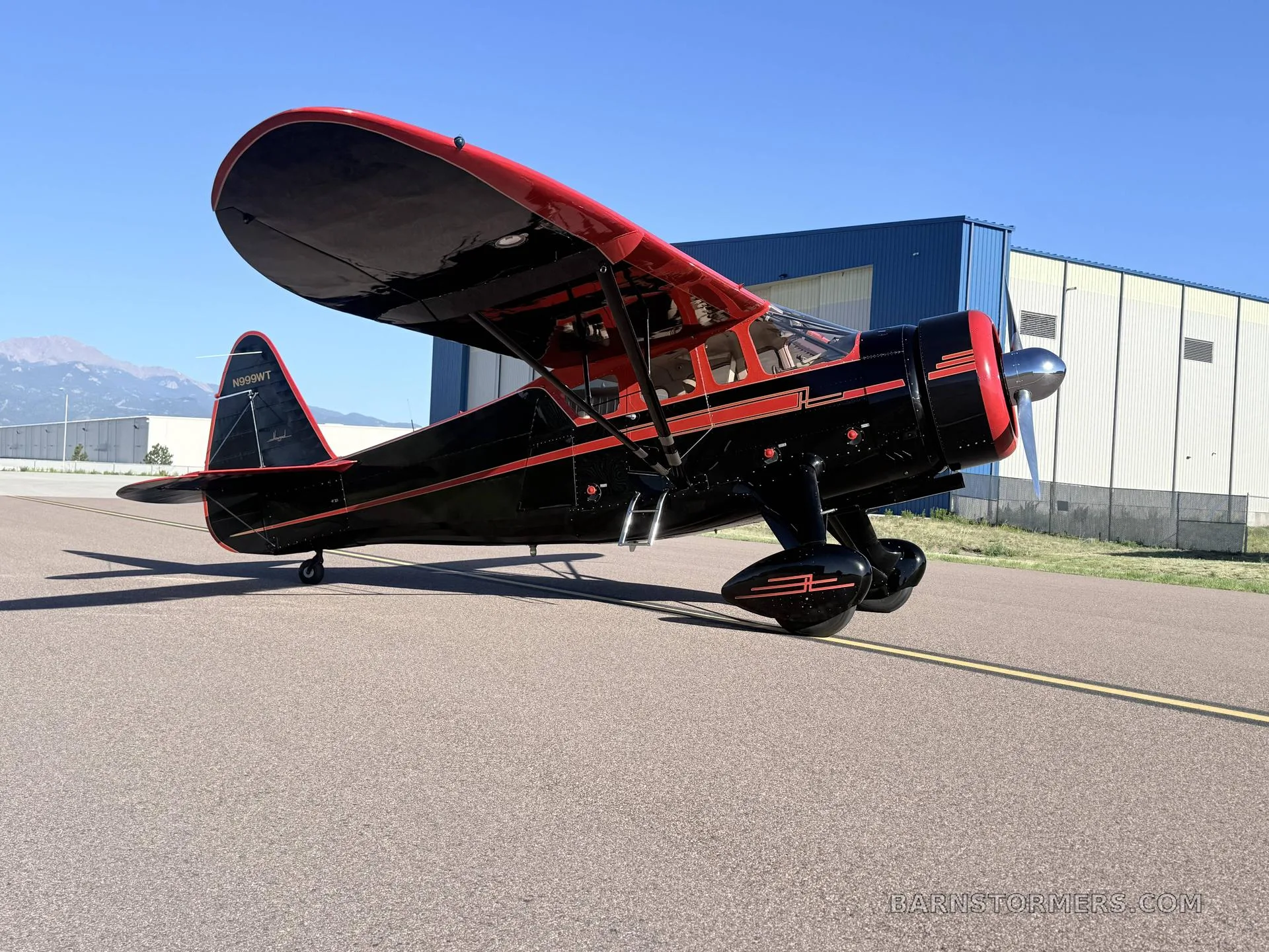 A black and red vintage high-wing monoplane with tail number N999WT, parked on an asphalt runway next to a blue hangar.