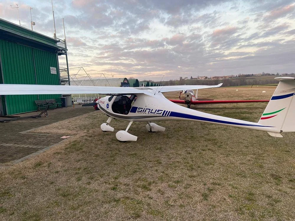 A white Pipistrel Sinus aircraft with blue and green tail stripes, parked on a grassy area next to a green hangar under a cloudy sky.