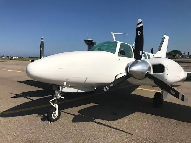 Front view of a white twin-engine propeller aircraft on an airport tarmac with a control tower in the background.