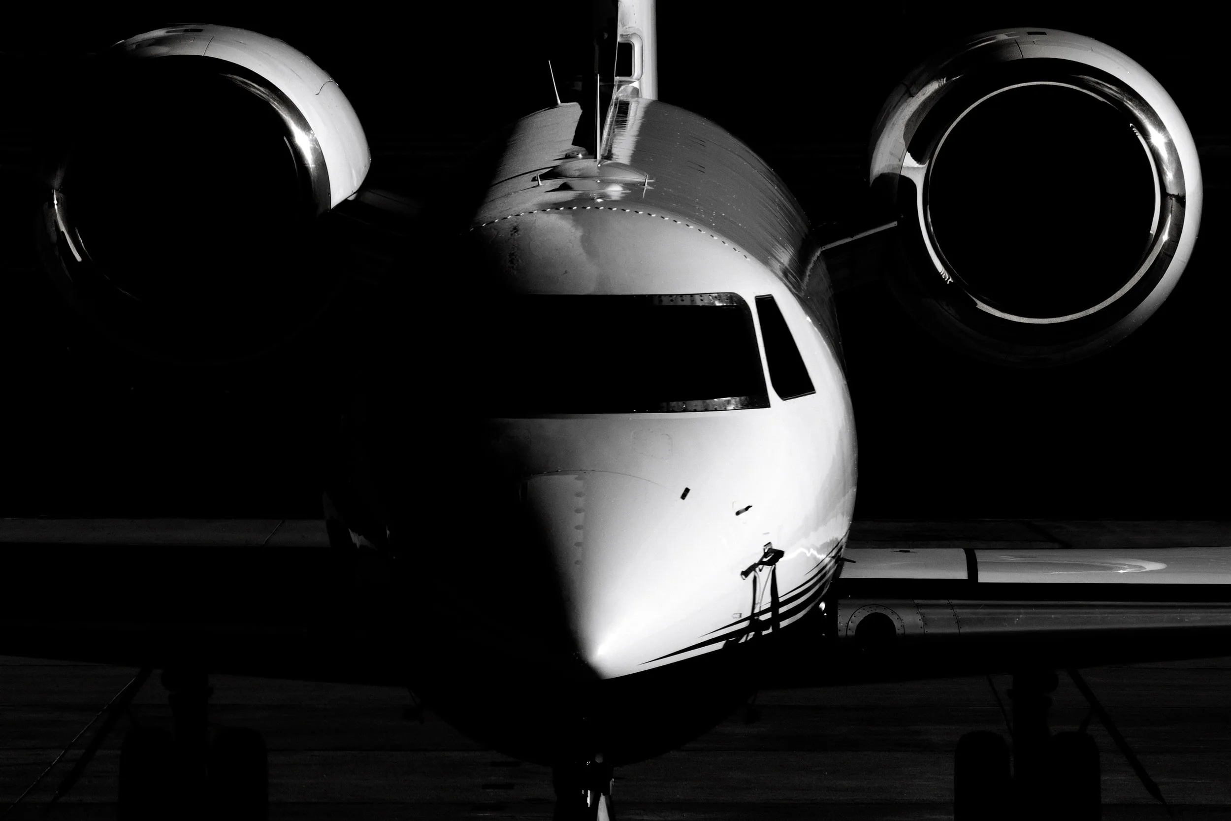 A black and white, head-on view of a business jet, highlighting its nose, cockpit, and engine nacelles against a dark background.