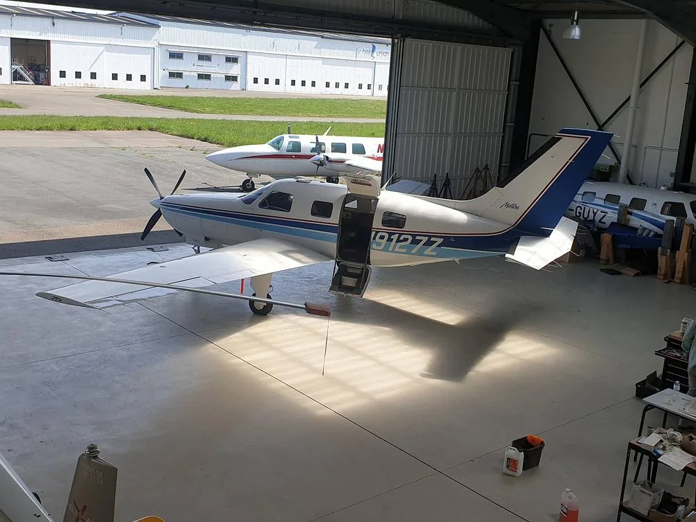 A white and blue Piper PA-46-350P Malibu Mirage aircraft, registration N9127Z, parked inside a hangar with its door open.